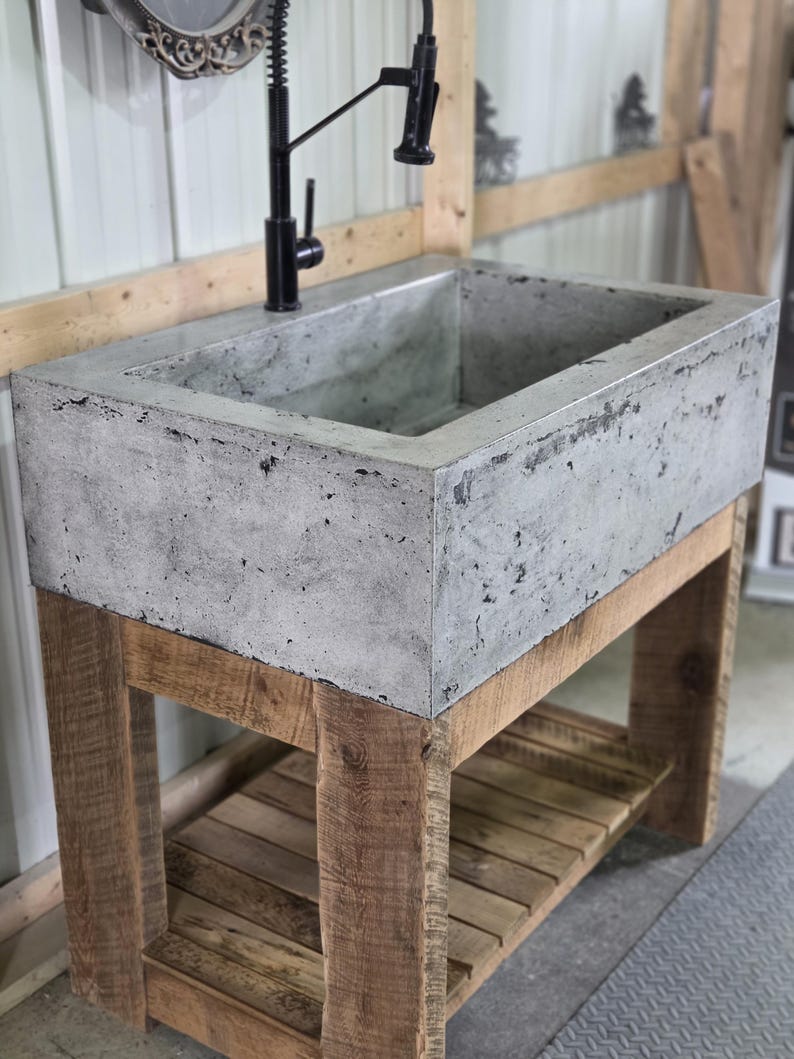 May include: A rectangular concrete farmhouse sink with a textured gray finish, mounted on a wooden vanity base with a slatted lower shelf. A tall, black gooseneck faucet with a pull-down sprayer is installed directly into the concrete basin.