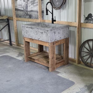 May include: A rectangular concrete sink basin with a mottled gray finish, mounted on a rustic wooden vanity base with four thick legs and a slatted lower shelf. A tall, black gooseneck faucet with a pull-down sprayer is installed on the sink.