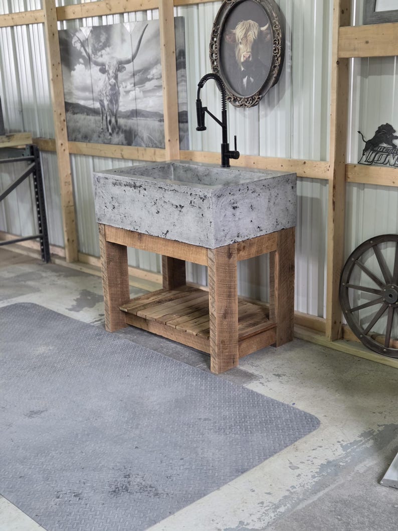 May include: A rectangular concrete sink basin with a mottled gray finish, mounted on a rustic wooden vanity base with four thick legs and a slatted lower shelf. A tall, black gooseneck faucet with a pull-down sprayer is installed on the sink.