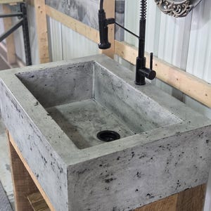 May include: A rectangular concrete sink with a mottled gray finish and a black drain, paired with a tall, black, industrial-style spring-neck faucet. The sink is mounted on a wooden base structure, showcasing a raw, textured aesthetic.