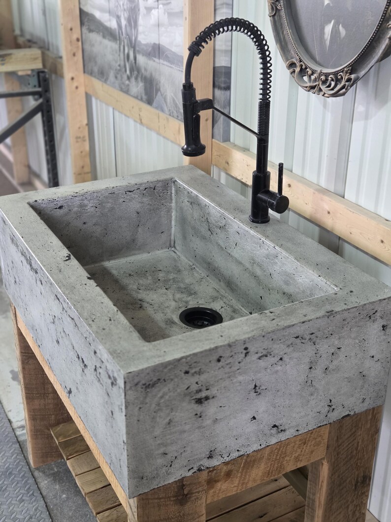 May include: A rectangular concrete sink with a mottled gray finish and a black drain, paired with a tall, black, industrial-style spring-neck faucet. The sink is mounted on a wooden base structure, showcasing a raw, textured aesthetic.
