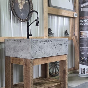 May include: A rectangular gray concrete sink basin mounted on a rustic wooden vanity base with a lower shelf. A tall black gooseneck faucet with a coiled spring neck is installed on the top surface of the sink. The vanity features four thick wooden legs.
