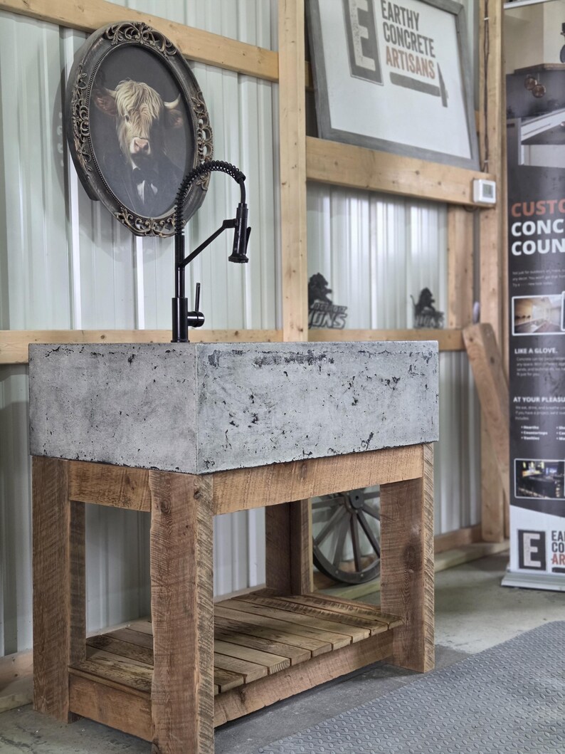 May include: A rectangular gray concrete sink basin mounted on a rustic wooden vanity base with a lower shelf. A tall black gooseneck faucet with a coiled spring neck is installed on the top surface of the sink. The vanity features four thick wooden legs.