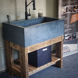 May include: A concrete sink with a black faucet is set on a reclaimed wood base. The sink is a dark gray color and has a rectangular shape. A black storage bin sits on the lower shelf. The sign says "CUSTOM CONCRETE COUNTERTOPS".