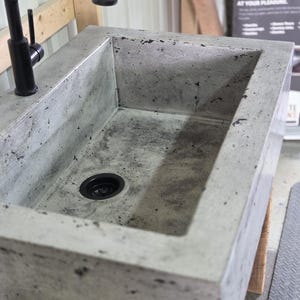 May include: A rectangular concrete sink with a mottled gray finish and a black drain. A tall, matte black faucet is mounted on the top edge of the sink. The basin features a textured surface with dark speckles throughout the material construction.