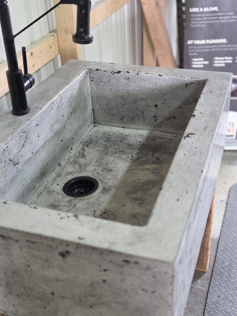 May include: A rectangular concrete sink with a mottled gray finish and a black drain. A tall, matte black faucet is mounted on the top edge of the sink. The basin features a textured surface with dark speckles throughout the material construction.