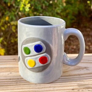 May include: A gray ceramic coffee mug with a raised design of a Super Nintendo controller. The controller has four buttons, one green, one blue, one yellow, and one red.