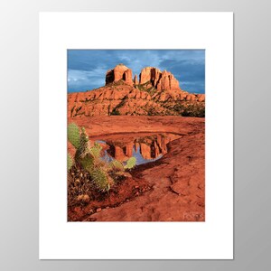 May include: A landscape photograph of a red rock formation under a blue sky. The formation is reflected in a small pool of water in the foreground. A green cactus is in the lower left corner.