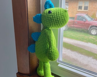May include: A green crocheted dinosaur toy with blue accents standing on a wooden windowsill. The dinosaur has a long neck, a small head, and a long tail.