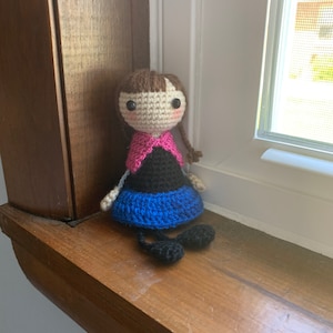 Peut inclure: Une poup&eacute;e en crochet avec des cheveux bruns, des v&ecirc;tements roses et noirs et des chaussures bleues. La poup&eacute;e est assise sur une surface en bois.