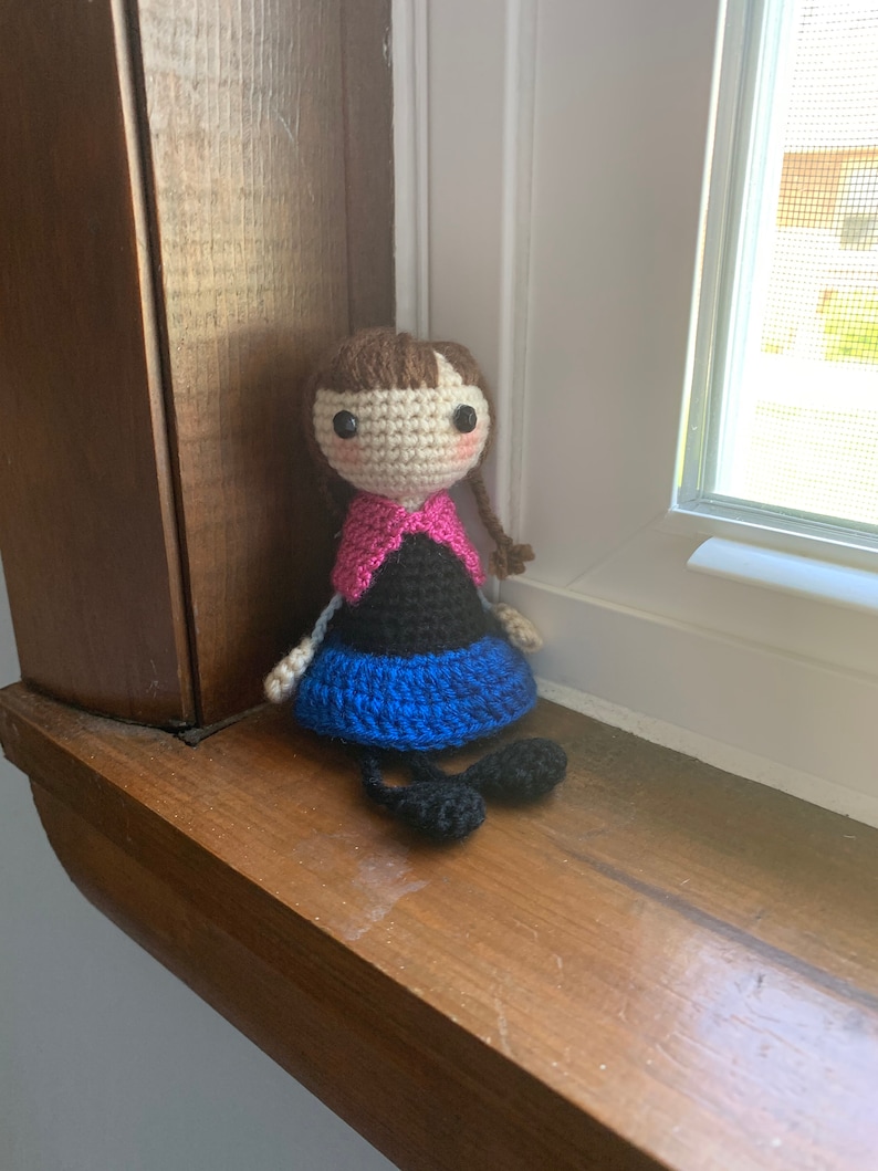 Peut inclure: Une poup&eacute;e en crochet avec des cheveux bruns, des v&ecirc;tements roses et noirs et des chaussures bleues. La poup&eacute;e est assise sur une surface en bois.