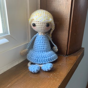 May include: A crocheted doll with blonde hair, blue dress, and blue shoes. The doll is sitting on a wooden surface.