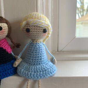 May include: A crocheted doll with blonde hair, a blue dress, and tan legs. The doll is sitting on a white windowsill.