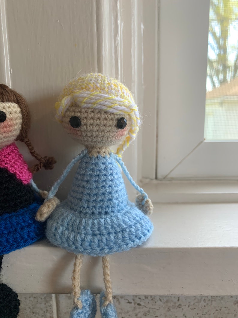 May include: A crocheted doll with blonde hair, a blue dress, and tan legs. The doll is sitting on a white windowsill.