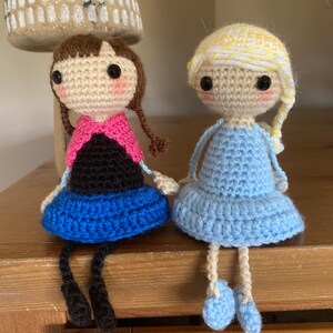 Peut inclure: Deux poup&eacute;es en crochet assises sur une surface en bois. La poup&eacute;e de gauche a les cheveux bruns, une &eacute;charpe rose, un haut noir et une jupe bleue. La poup&eacute;e de droite a les cheveux blonds, une robe bleue et des chaussures bleues.