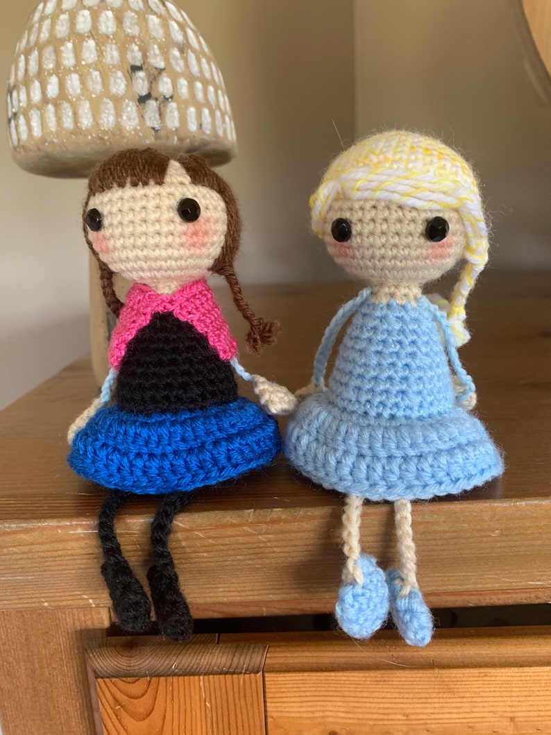 Peut inclure: Deux poup&eacute;es en crochet assises sur une surface en bois. La poup&eacute;e de gauche a les cheveux bruns, une &eacute;charpe rose, un haut noir et une jupe bleue. La poup&eacute;e de droite a les cheveux blonds, une robe bleue et des chaussures bleues.