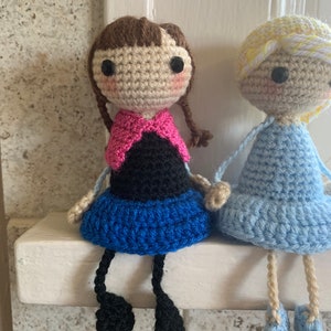 Peut inclure: Deux poup&eacute;es en crochet assises sur une &eacute;tag&egrave;re blanche. La poup&eacute;e de gauche a les cheveux bruns, un haut rose et noir, une jupe bleue et des jambes noires. La poup&eacute;e de droite a les cheveux blonds, une robe bleu clair et des jambes beiges.