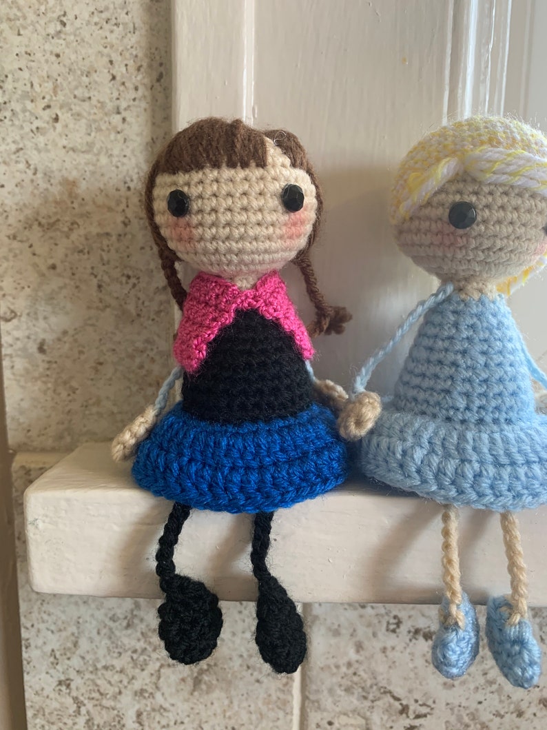 Peut inclure: Deux poup&eacute;es en crochet assises sur une &eacute;tag&egrave;re blanche. La poup&eacute;e de gauche a les cheveux bruns, un haut rose et noir, une jupe bleue et des jambes noires. La poup&eacute;e de droite a les cheveux blonds, une robe bleu clair et des jambes beiges.