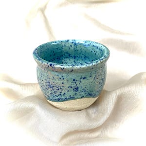 May include: A small, handmade ceramic pot with a speckled turquoise glaze. The pot has a rounded shape with a slightly flared rim and a natural, unglazed base. Dark blue speckles add detail to the turquoise color.
