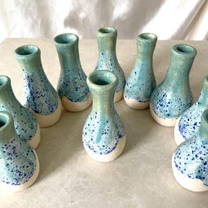 May include: A collection of ten small ceramic bud vases arranged in a group. Each vase features a narrow neck and a rounded base with a speckled blue and teal glaze finish that transitions into a solid, unglazed cream-colored bottom section.