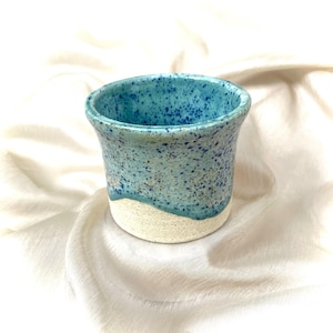 May include: A small, handmade ceramic tumbler with a speckled turquoise glaze. The top and sides are a vibrant turquoise with darker blue speckles, transitioning to a creamy white base. The cup has a slightly flared rim.