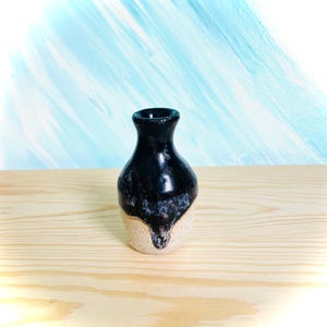 May include: A small ceramic vase with a narrow neck and a bulbous body. The upper portion features a glossy black glaze that drips down over the lower section, which has a textured, matte, off-white finish. The vase has a simple, rounded rim.