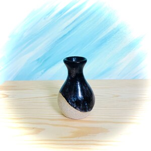 May include: A small ceramic vase with a bulbous base and a narrow neck. The upper portion of the vase features a glossy, dark black glaze, while the lower portion displays a textured, unglazed beige surface with a visible speckled finish.