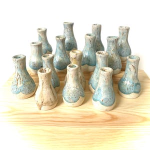 May include: A collection of small, handmade ceramic vases. Each vase features a unique design with a light blue and cream color palette. The vases are arranged on a light-colored wooden surface, showcasing their individual shapes and textures.