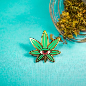 May include: A gold-coloured pin featuring a green marijuana leaf design. The leaf has a red eye in the centre. The pin is on a turquoise background.