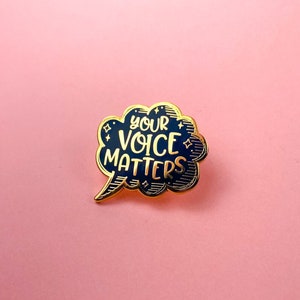 May include: Gold enamel pin with a black speech bubble design. The speech bubble says "Your Voice Matters".