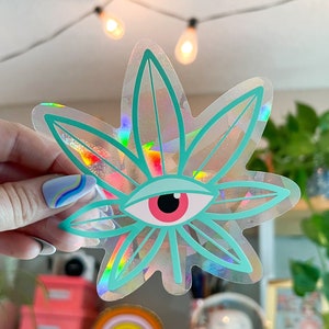 K&ouml;nnte beinhalten: Ein holografischer Aufkleber mit einem gr&uuml;nen Marihuanablatt und einem roten Auge in der Mitte.