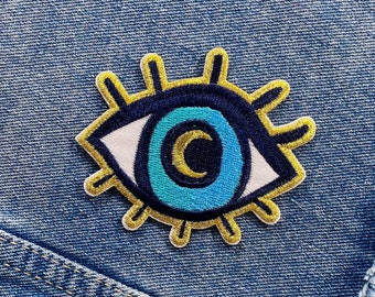 Puede incluir: Un ojo azul y blanco con una luna creciente dorada en su interior, rodeado de un sol radiante dorado, está bordado en una tela vaquera azul.