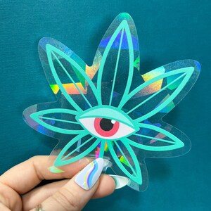 K&ouml;nnte beinhalten: Ein holografischer Aufkleber mit einem gr&uuml;nen Marihuanablatt und einem gro&szlig;en roten Auge in der Mitte. Der Aufkleber hat einen regenbogenfarbenen, irisierenden &Uuml;berzug.
