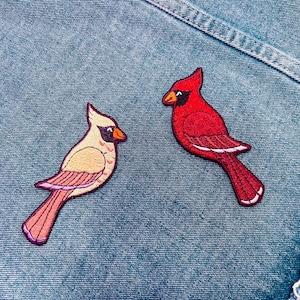 May include: Two embroidered cardinal bird patches. One cardinal is red with black and white accents, the other is tan with pink and purple accents. The patches are on a blue denim background.