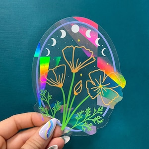 May include: A holographic sticker featuring a floral design with three flowers and leaves. The flowers are outlined in gold and the background is a rainbow gradient with a crescent moon pattern.