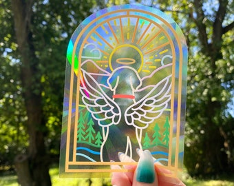 Puede incluir: Una pegatina holográfica con un fondo de arcoíris y un contorno blanco de un perro con alas de ángel. El perro lleva un halo y un collar. La pegatina está en un estilo de vitral.