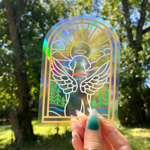 May include: A holographic sticker with a rainbow background and a white outline of a dog with angel wings. The dog is wearing a halo and a collar. The sticker is in a stained glass style.