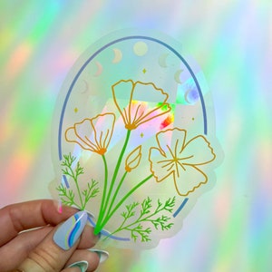 May include: A clear sticker with a holographic finish featuring a stylised illustration of a bouquet of yellow and white flowers with green leaves. The flowers are surrounded by a blue circle with a crescent moon design.
