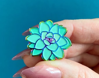 Puede incluir: Un pin de esmalte verde e iridiscente con forma de suculenta con un solo ojo morado en el centro.