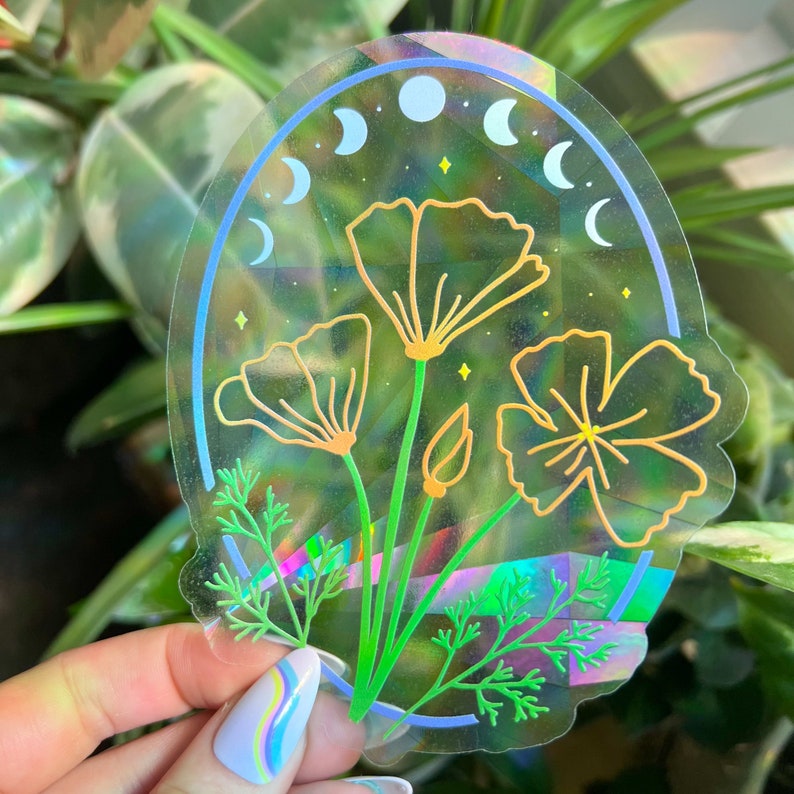 May include: A holographic sticker featuring a cluster of yellow and orange flowers with green stems and leaves. The sticker has a blue and white crescent moon design around the flowers.