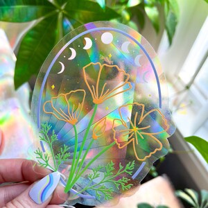 May include: A holographic sticker with a rainbow background and a design of three orange flowers with green stems and leaves. The sticker also features a blue crescent moon design.