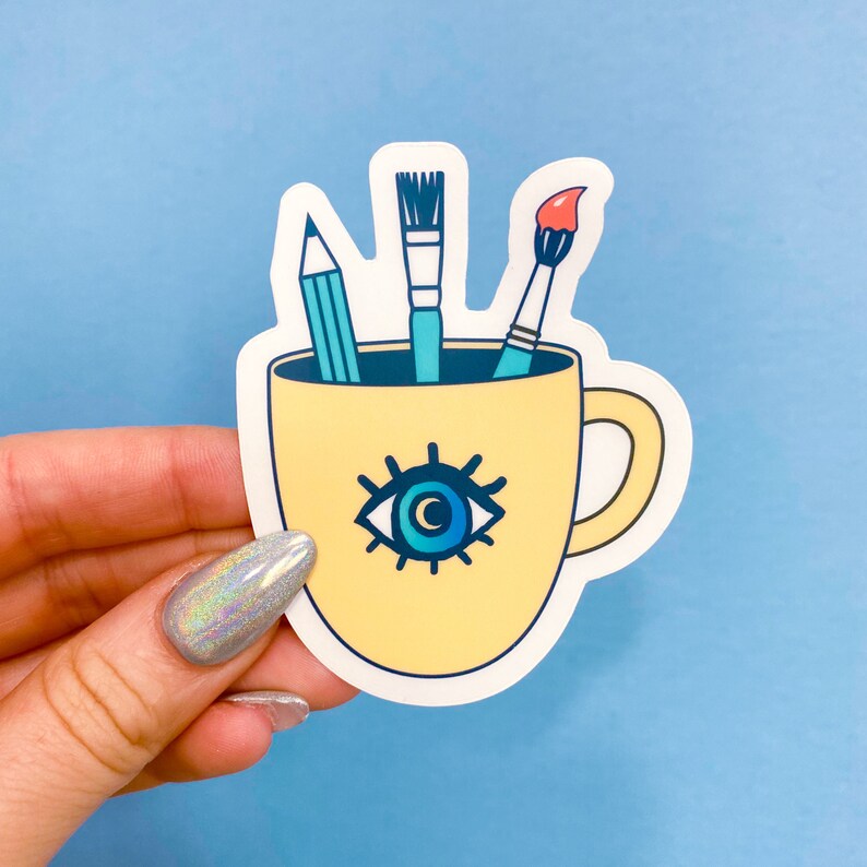 Puede incluir: Una taza de cer&aacute;mica amarilla con un dise&ntilde;o de ojo azul y un asa. Tres pinceles sobresalen de la taza, uno azul, uno rojo y uno amarillo.