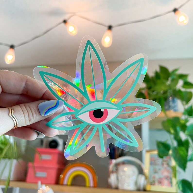 K&ouml;nnte beinhalten: Ein holografischer Aufkleber mit einem gr&uuml;nen Marihuanablatt und einem roten Auge in der Mitte.