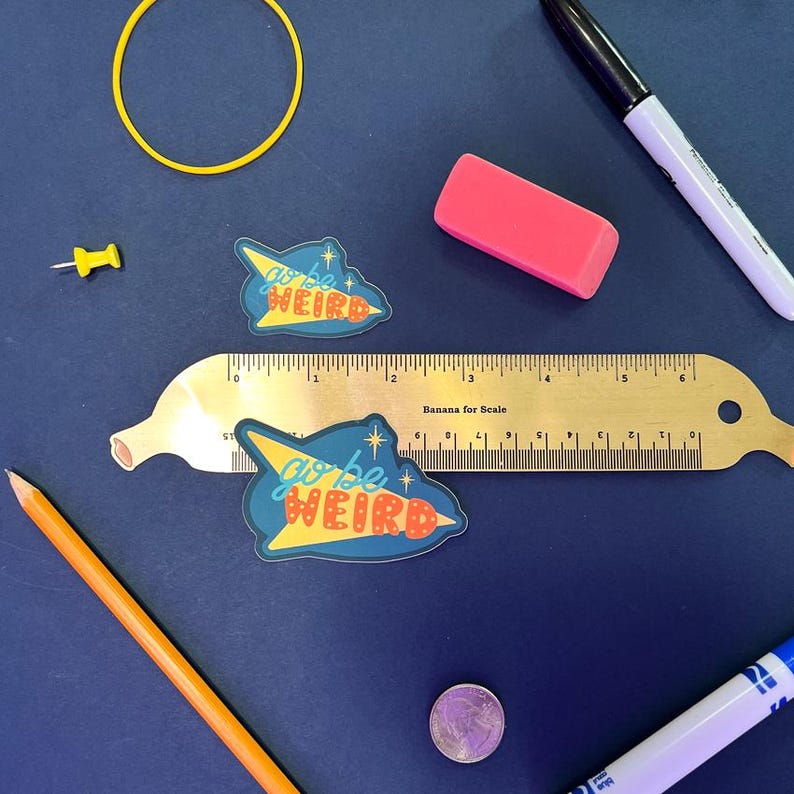 Puede incluir: Un surtido de &uacute;tiles escolares y de oficina sobre una superficie azul oscuro. Incluye una regla dorada, dos pegatinas con el texto "go be weird", una goma de borrar rosa, un rotulador negro, un chincheta amarilla, una goma el&aacute;stica amarilla, un l&aacute;piz y una moneda.