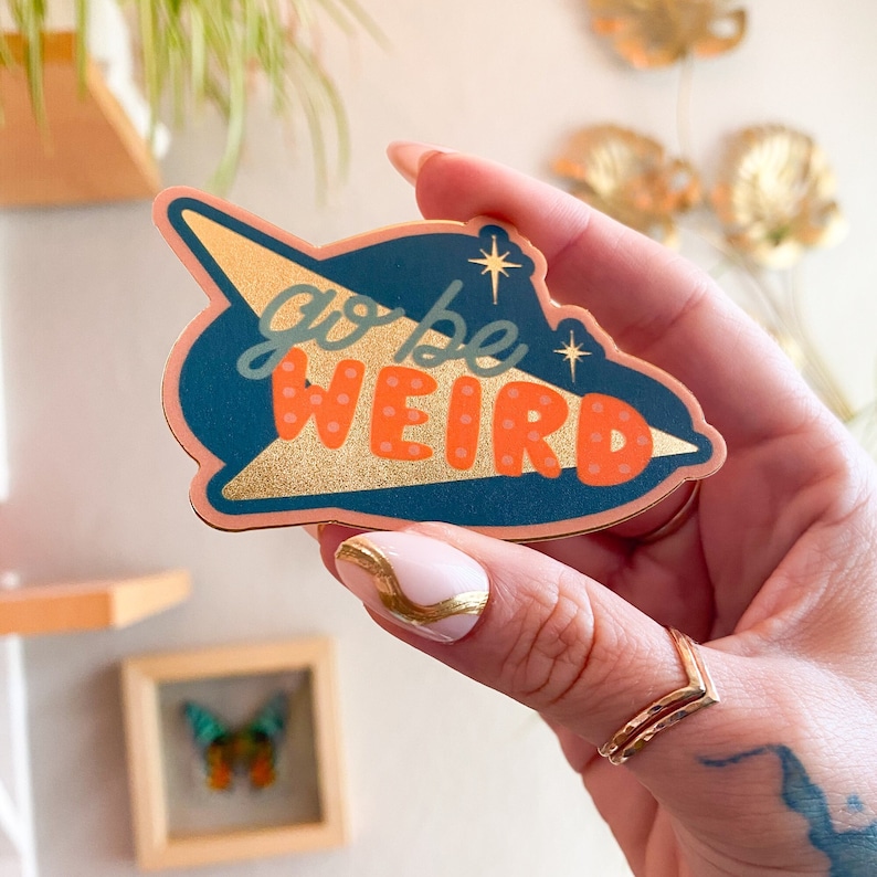 Puede incluir: Una pegatina con un dise&ntilde;o retro, con las palabras "go be WEIRD" en verde azulado y naranja. La pegatina tiene un acento de l&aacute;mina dorada y una combinaci&oacute;n de colores verde azulado y melocot&oacute;n. La pegatina tiene forma de cohete retro.