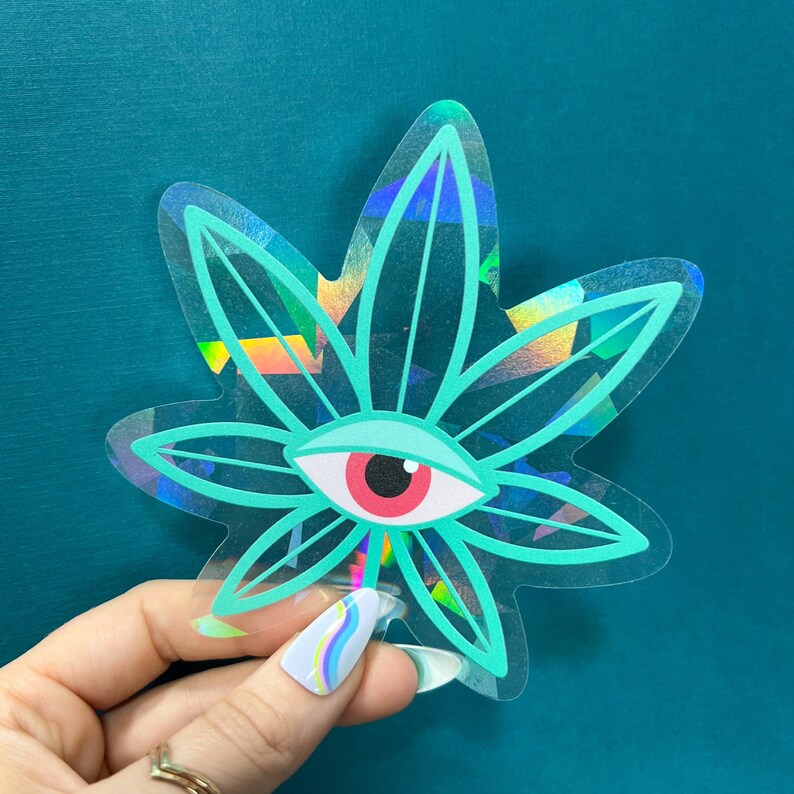 K&ouml;nnte beinhalten: Ein holografischer Aufkleber mit einem gr&uuml;nen Marihuanablatt und einem gro&szlig;en roten Auge in der Mitte. Der Aufkleber hat einen regenbogenfarbenen, irisierenden &Uuml;berzug.