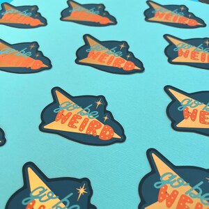 Puede incluir: Una colecci&oacute;n de pegatinas sobre un fondo turquesa. Cada pegatina presenta una forma azul oscuro con un dise&ntilde;o amarillo y naranja y las palabras "go be WEIRD" en naranja y blanco. Las pegatinas tienen una est&eacute;tica retro y espacial.