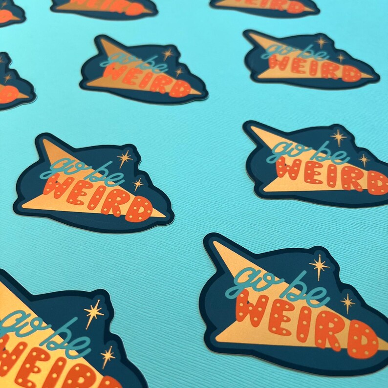 Puede incluir: Una colecci&oacute;n de pegatinas con forma de nube azul marino, una flecha amarilla y las palabras "go be WEIRD" en naranja. Las pegatinas est&aacute;n dispersas sobre una superficie azul claro.