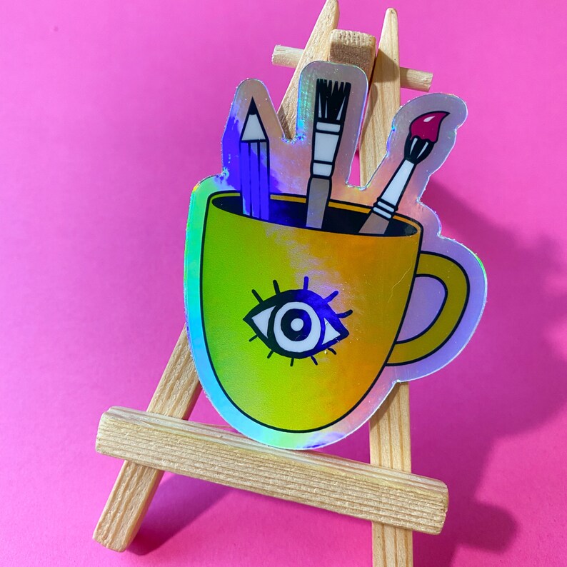 Puede incluir: Una pegatina hologr&aacute;fica de una taza amarilla con un dise&ntilde;o de ojo y tres &uacute;tiles de arte que sobresalen de la parte superior. Los &uacute;tiles de arte son un l&aacute;piz, un pincel y un pincel con una punta roja.