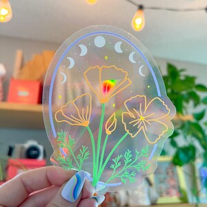 May include: A clear sticker with a holographic finish featuring a design of three orange California poppies with green stems and leaves. The background is a blue and white crescent moon design.